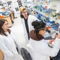 Two students talking to a third student sitting at a microscope