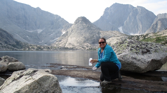 Leora Nanus kneeling at alpine lake surrounded by mountains